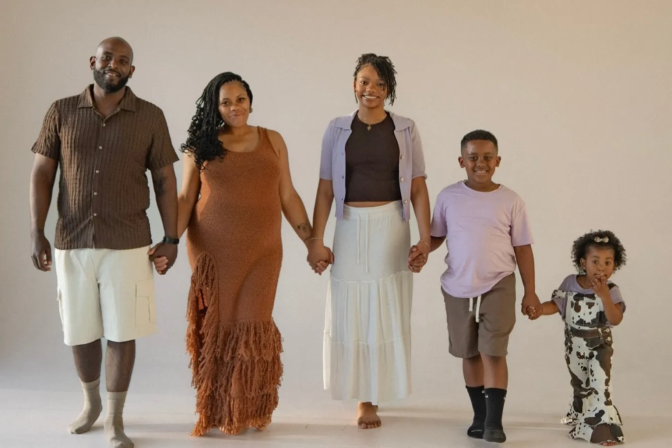 A family of five walking hand in hand across a bright, empty studio, dressed in earth tones and soft lavender.